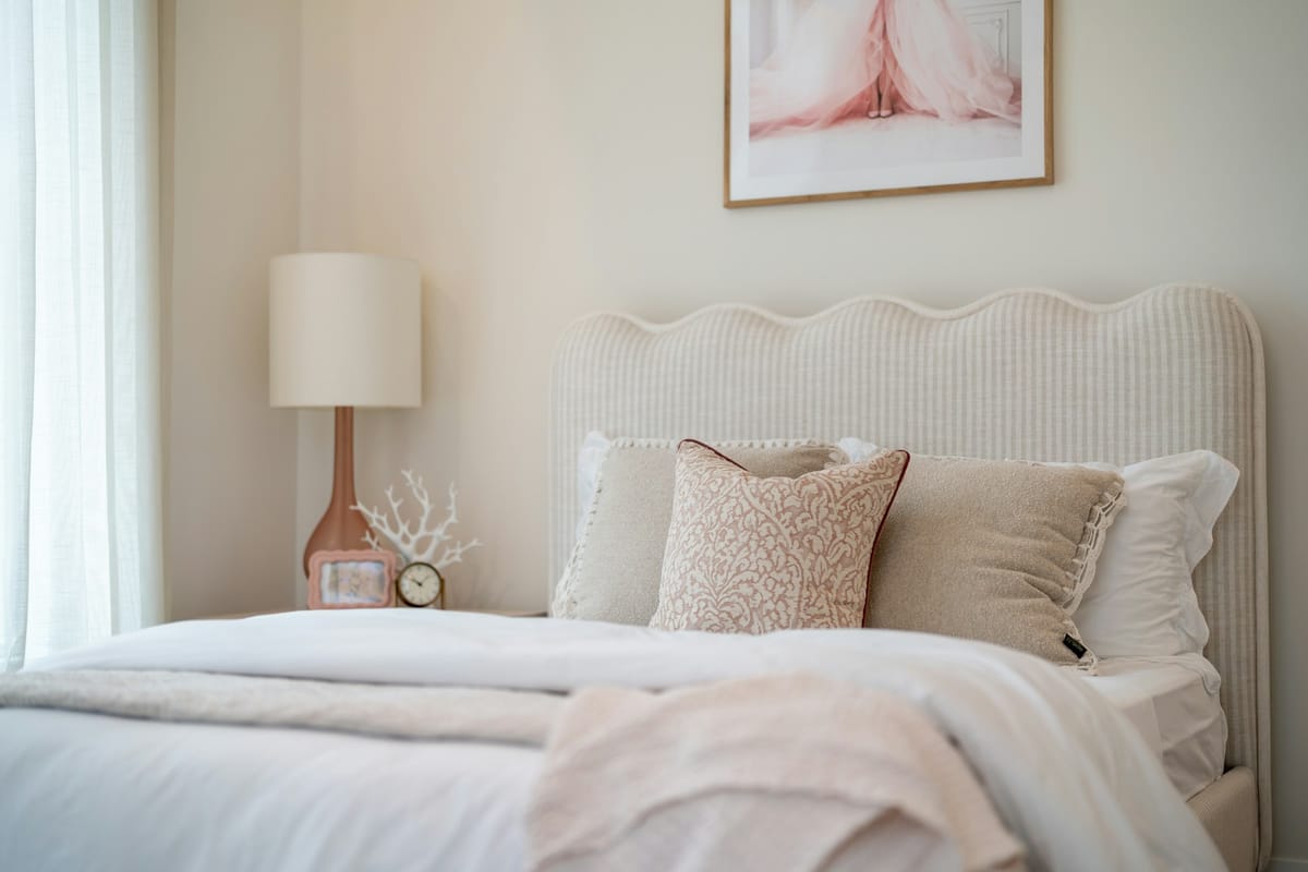A soft white bed with a wavy headboard, blush pillows, and a bedside lamp